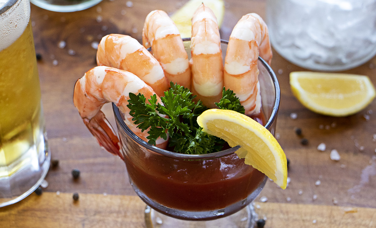 A clear drink cup filled with cocktail juice sauce, parsley, bay leafs, shrimp, and lemon