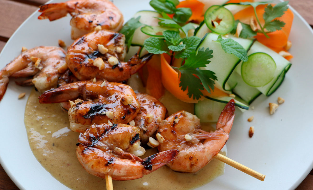 A plate of shrimp on 2 wooden skewers with coconut green thai curry sauce and cucumber & carrots.